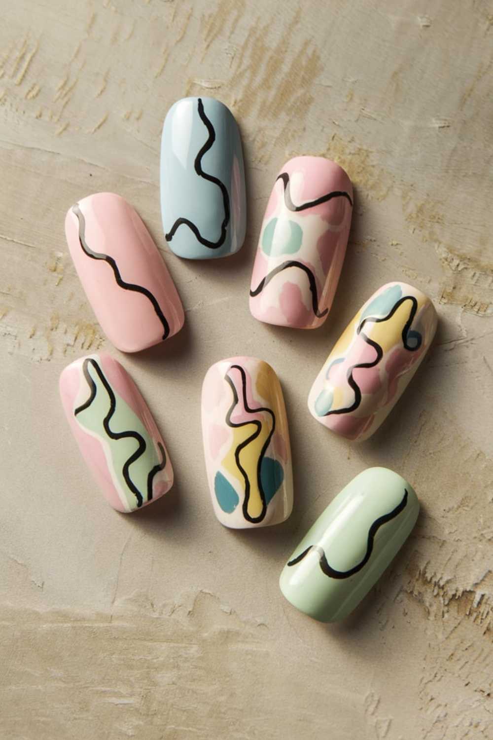 A set of seven artificial nails on a textured surface showcases simple nail art with abstract pastel designs and wavy black lines. Colors include light pink, mint green, soft yellow, pale blue, and lavender.