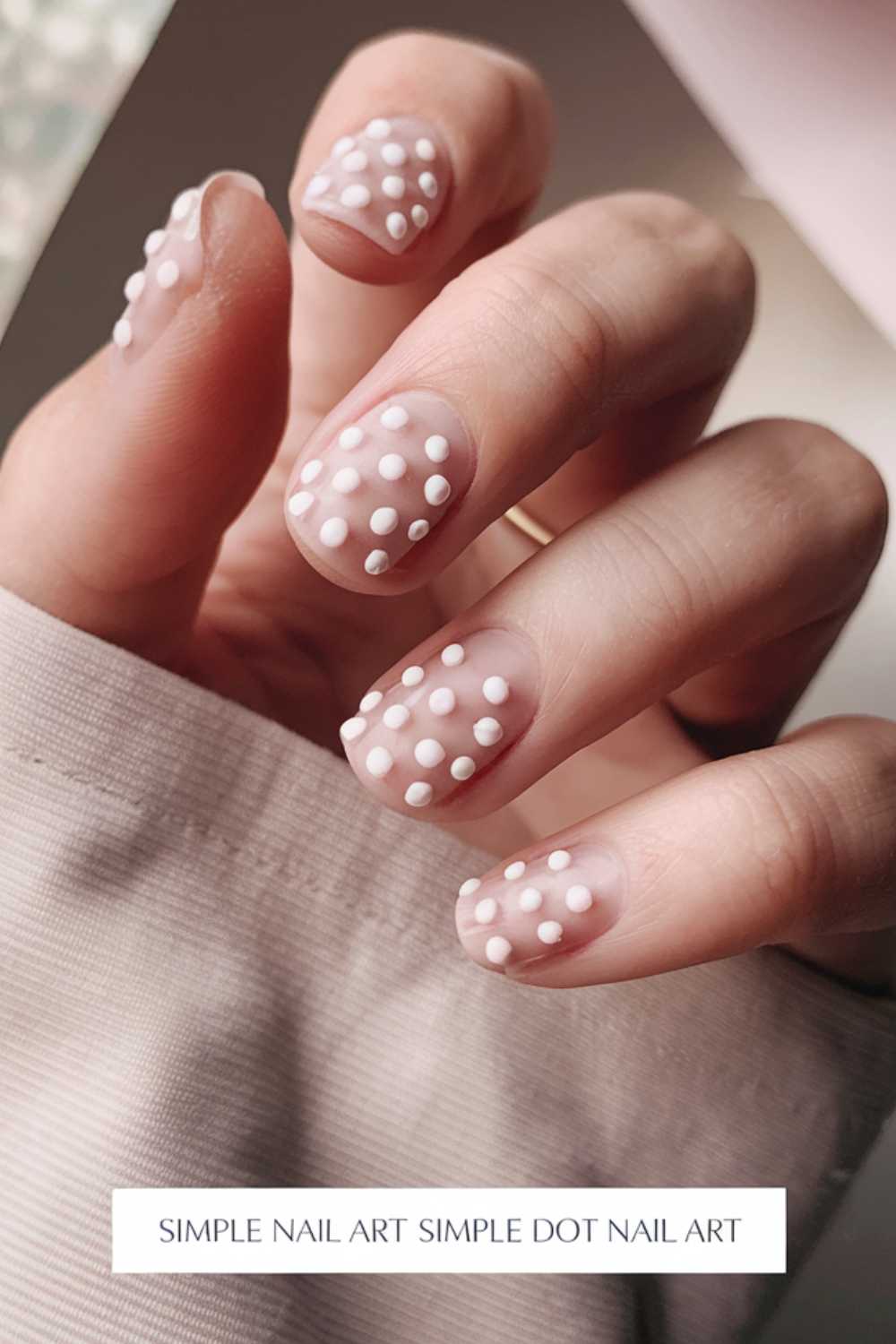 A close-up showcases a hand with nails painted in light nude, adorned with delicate white polka dots. Slightly bent fingers peek from a beige sleeve, embodying the essence of Simple Nail Art. Text reads Simple Dot Nail Art.