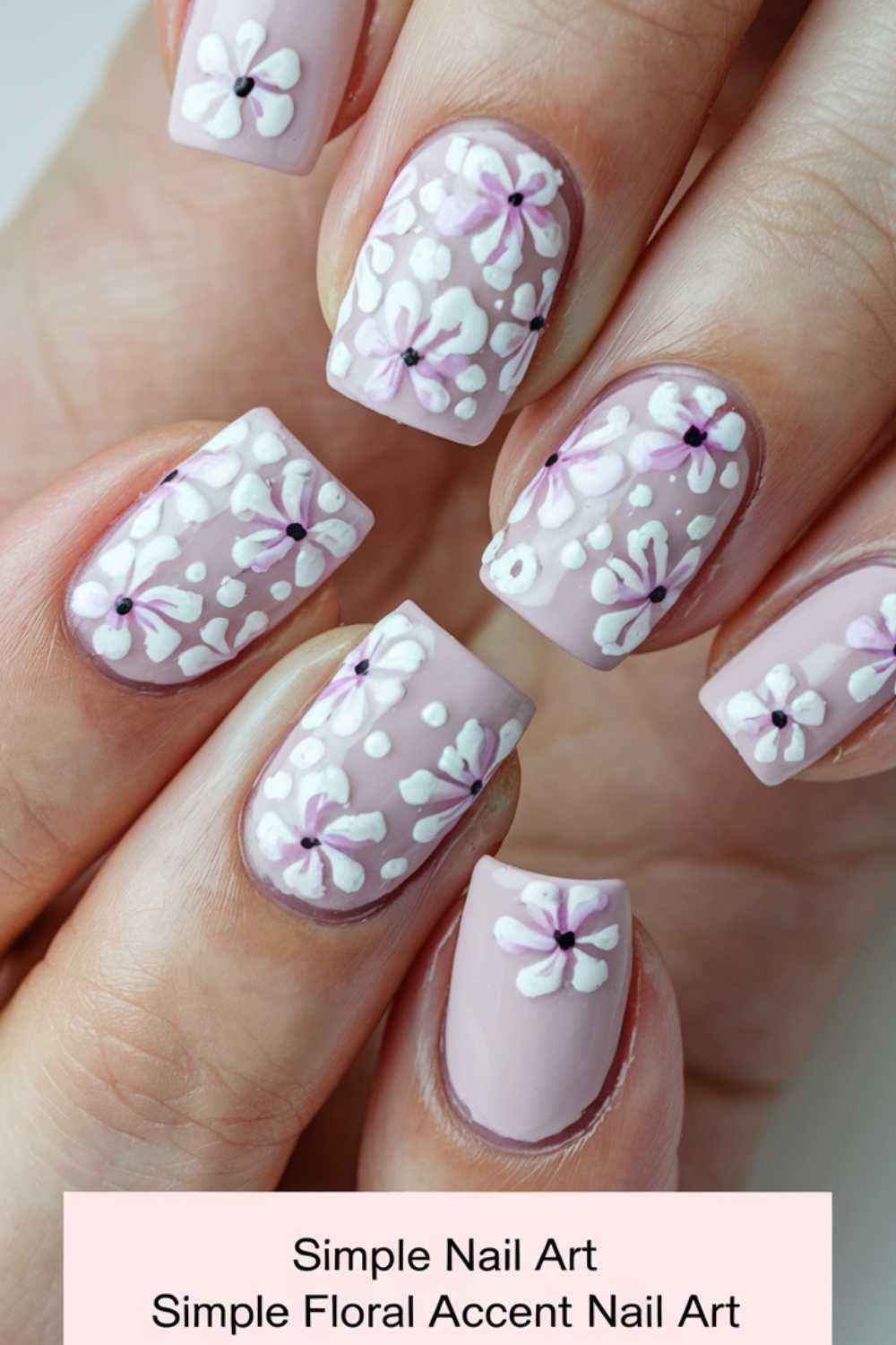 Close-up of hands adorned with light pink polish and featuring delicate white and purple floral designs on most nails. Titled Simple Nail Art, this image elegantly showcases the charm of simple floral accent nail art.