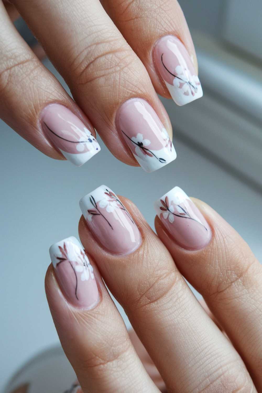 Close-up of manicured hands showcasing elegant, simple nail art featuring French tips and delicate floral designs. The nails boast a pink base with white tips, adorned with small white flowers and thin black branches for a timeless look.