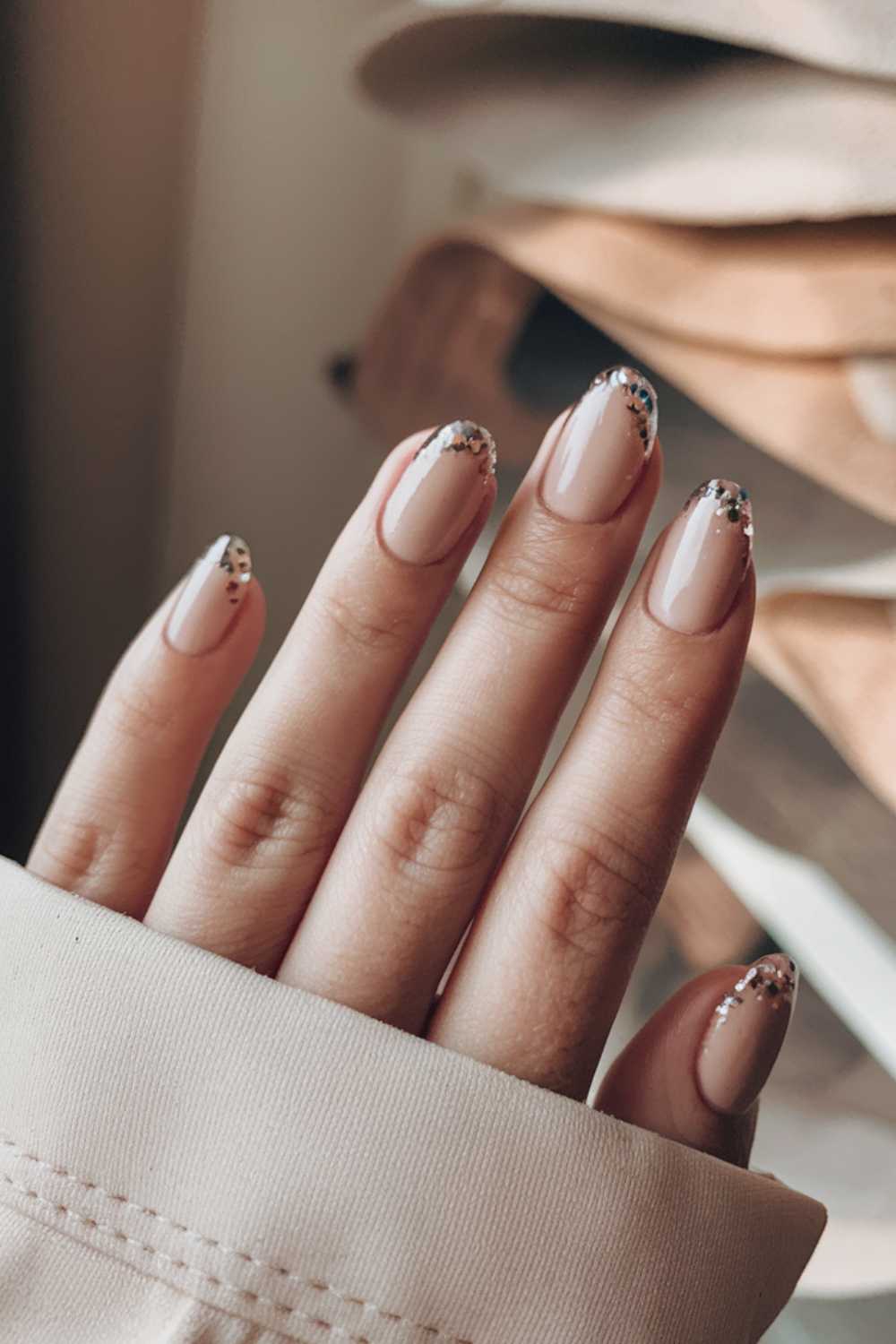 A hand with neatly manicured nails is shown, showcasing simple nail art. The nails have a glossy nude base color with a silver glitter gradient at the tips. The sleeve of a beige garment partially covers the wrist while the background remains softly blurred.