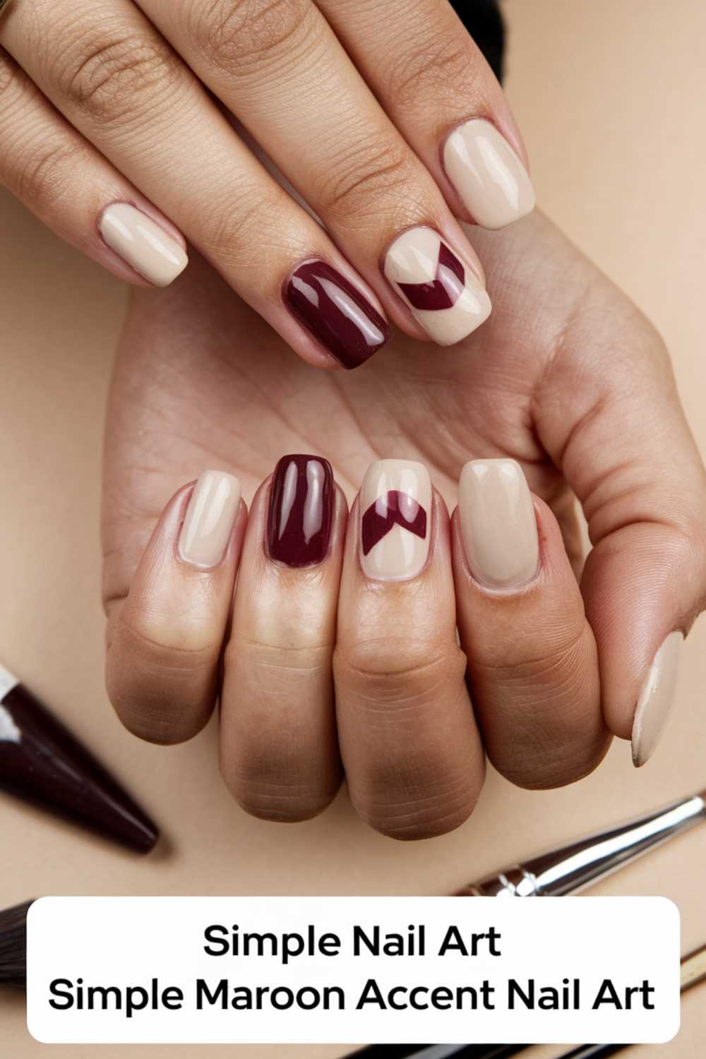 Close-up of a hand with a nude manicure featuring maroon accent designs. Some nails showcase solid maroon polish, while others sport elegant maroon tips or chevrons. Brushes lie beside the hand, emphasizing the Simple Nail Art theme with stylish maroon accents.