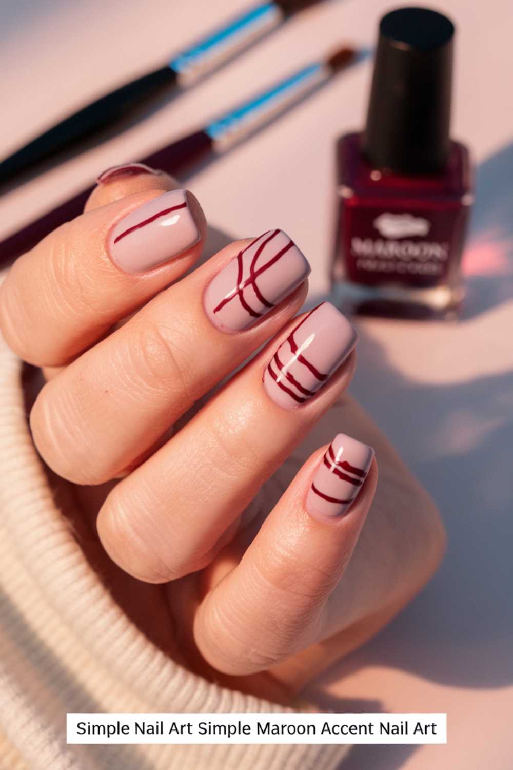 Close-up of a hand with pale pink nails showcasing simple nail art in maroon abstract lines. A maroon nail polish bottle and brushes are blurred in the background, while the person is wearing a cream-colored sweater.