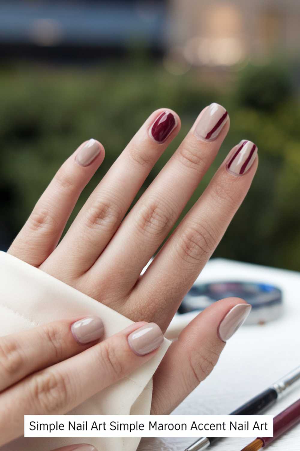 A hand showcasing simple nail art. Most nails are painted a smooth, beige color, while the ring finger features a bold, dark maroon accent with a glossy finish. The background is blurred, highlighting the elegant design.