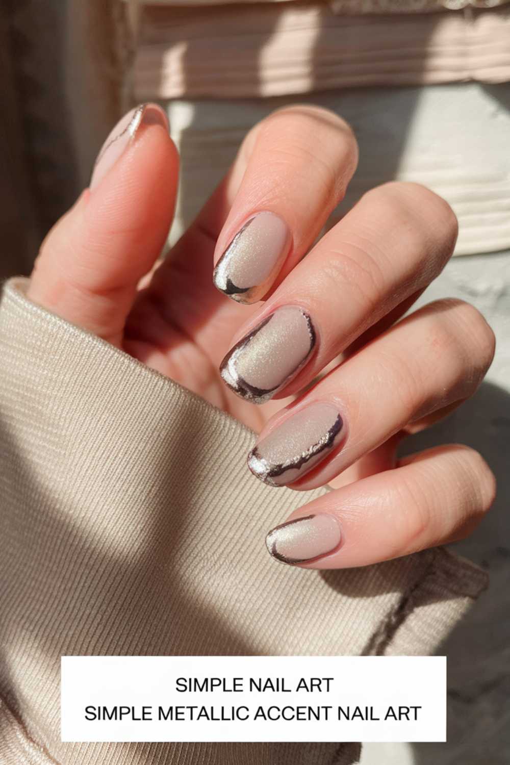 A hand showcases simple metallic accent nail art on a beige matte base, with shiny silver and dark metallic designs gracing the tips. A light beige sweater sleeve peeks into view. Text reads: Simple Nail Art - Effortlessly Elegant.