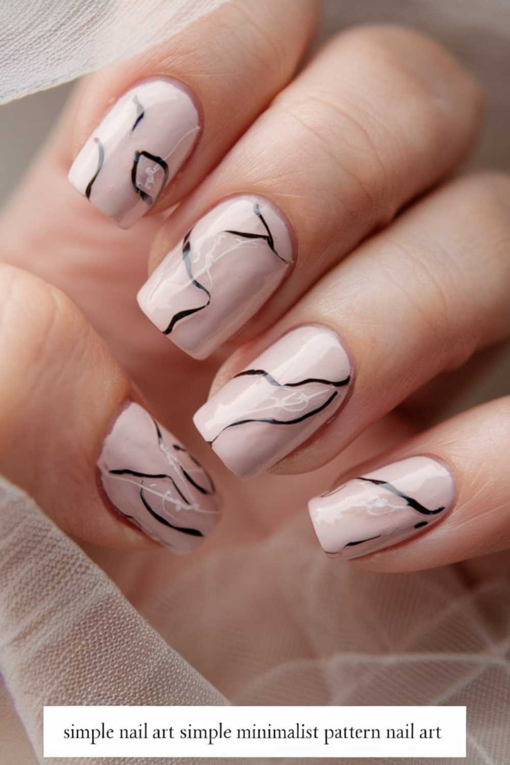 Close-up of a hand showcasing simple nail art with nails painted in a pale pink shade, adorned with minimalist black abstract line design. The skin is smooth, and a sheer white fabric drapes softly around the fingers.