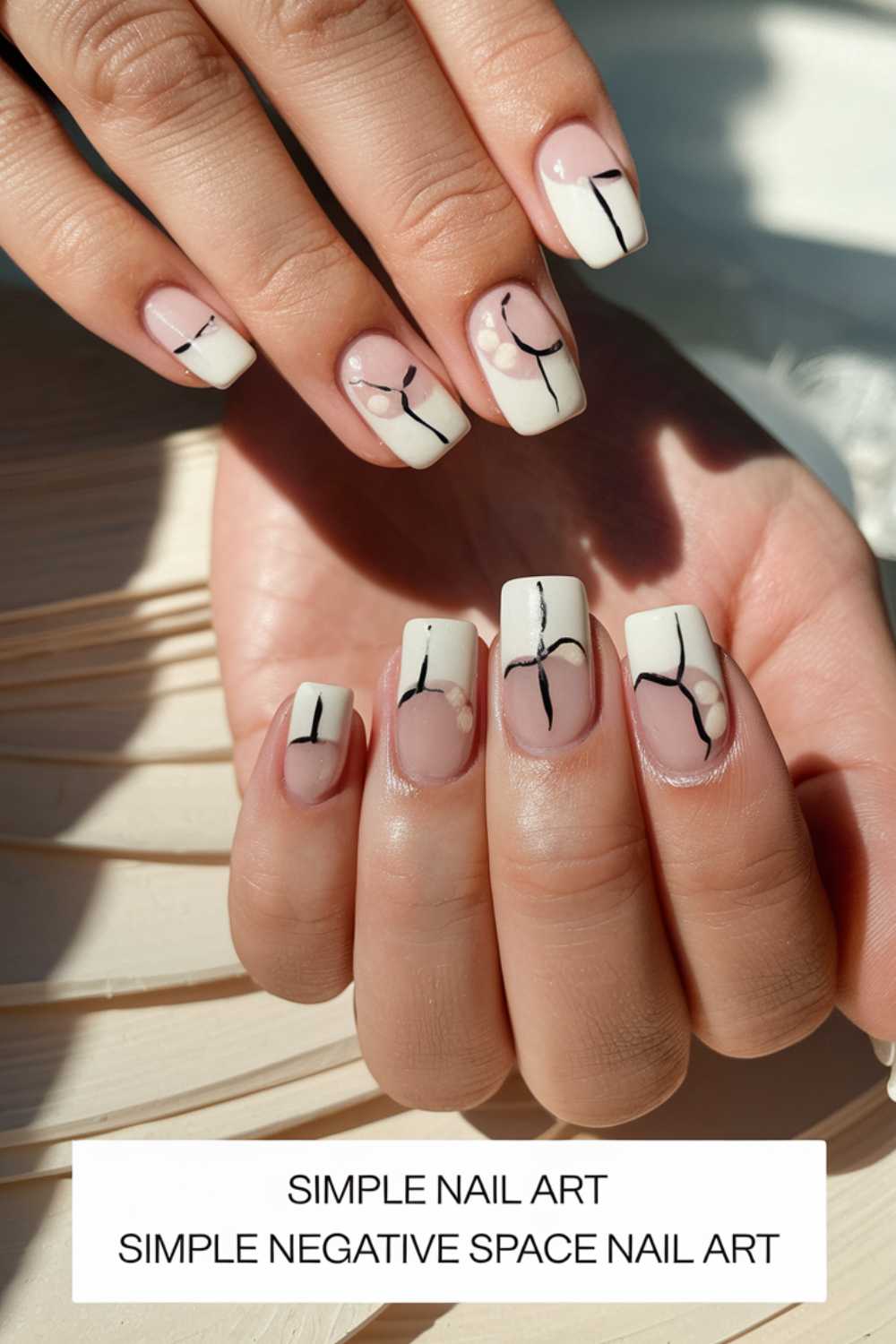 Close-up of hands with square-shaped nails showcasing a white and clear negative space design. Black abstract lines and dots adorn each nail, all beautifully captured against a beige background. Sunlight creates soft shadows, highlighting the elegance of this simple nail art.