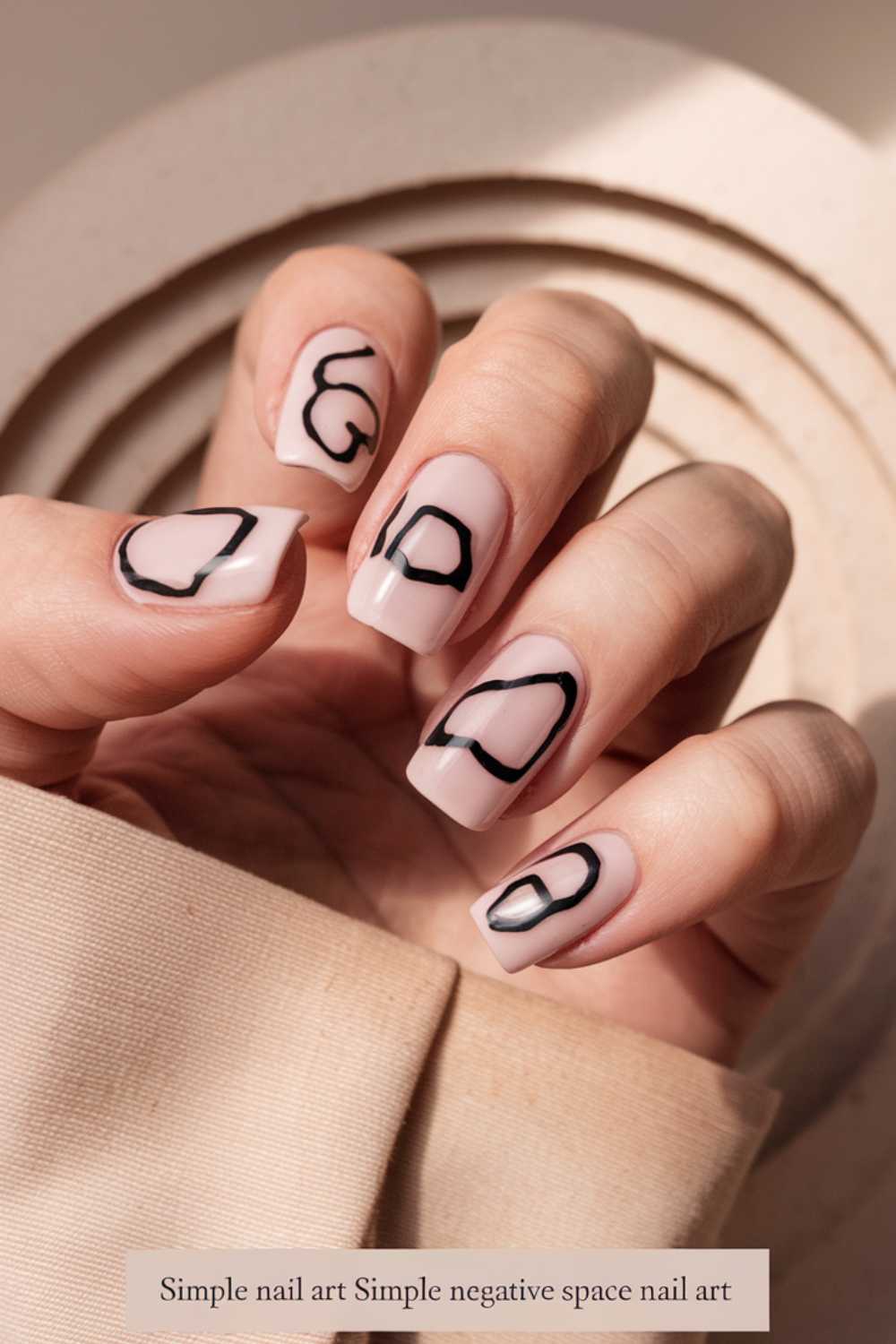Close-up of a hand showcasing simple nail art with neutral tones and abstract black line designs. The nails have a glossy finish and feature a modern, negative space style. A beige fabric is partially visible in the background, adding elegance to the look.