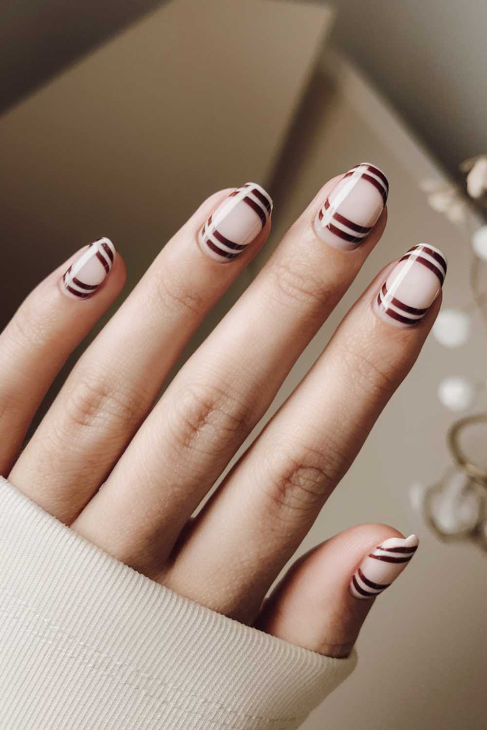 A hand with light skin is shown wearing a beige sleeve, adorned with simple nail art. The fingernails feature a creative design of burgundy stripes on a nude base. The background is blurred and neutral-toned.