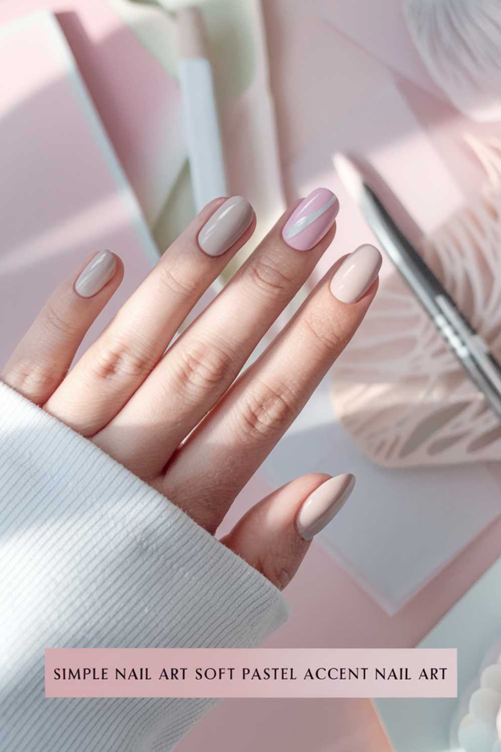A hand with long, oval-shaped nails showcases simple nail art in pastel pink and beige. One nail features a diagonal stripe design. The hand rests on aesthetically arranged pink and beige papers, overlaid with text that reads Simple Nail Art Soft Pastel Accent.