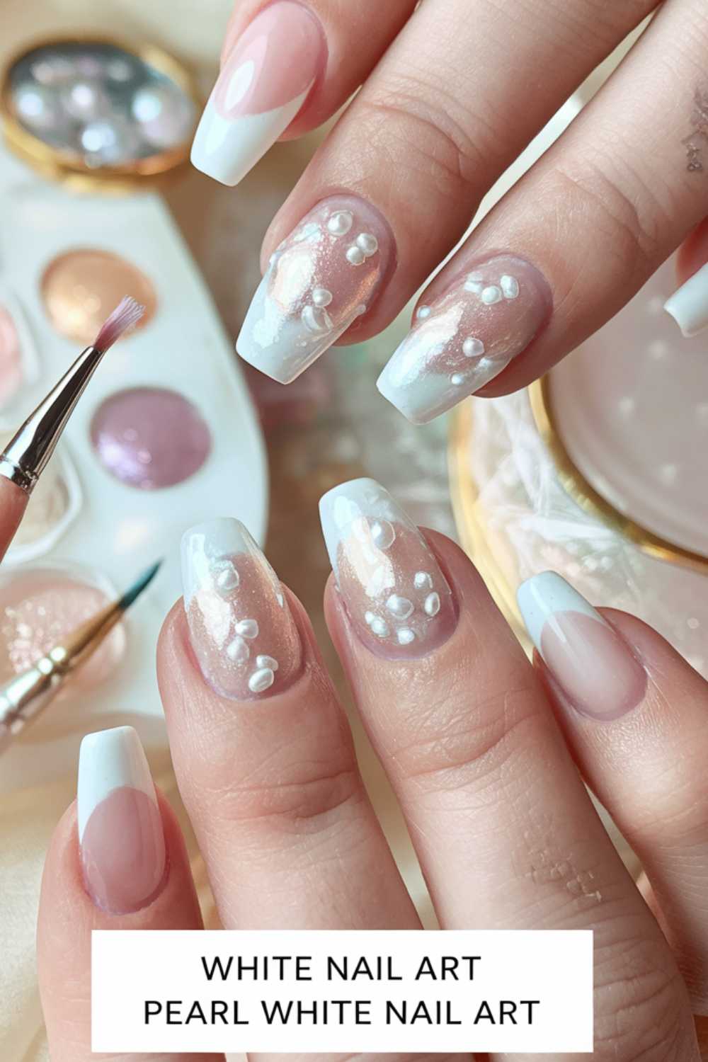 Close-up of hands showcasing French manicure with white tips and pearl accents. A brush is applying polish. Background includes nail art supplies and a small palette.