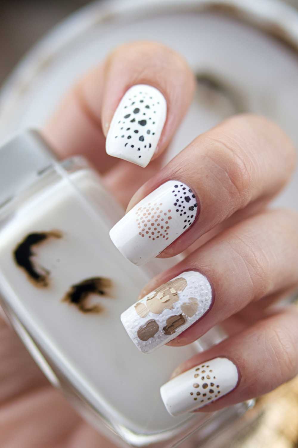 A hand displaying artistic nail art featuring white polish with abstract black and beige dot patterns on the nails. The hand is holding a bottle with a similar design theme.