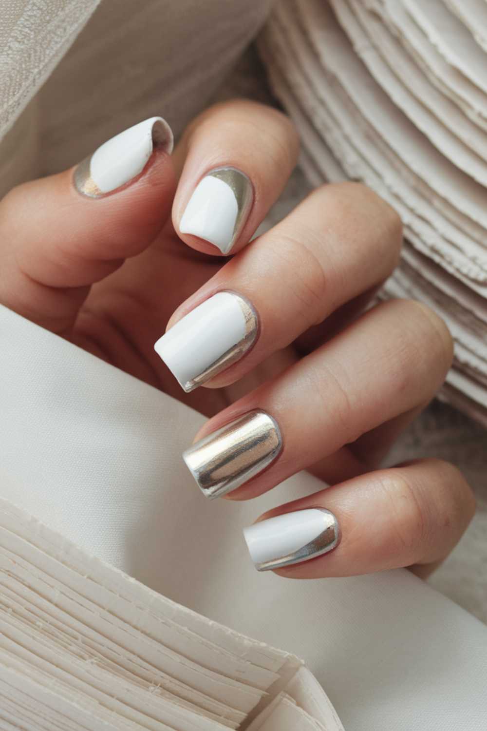 Close-up of a hand with a manicure featuring white and metallic silver nail polish on squared nails. The fingers rest against a white fabric and layered paper background, accentuating the elegant nail design.