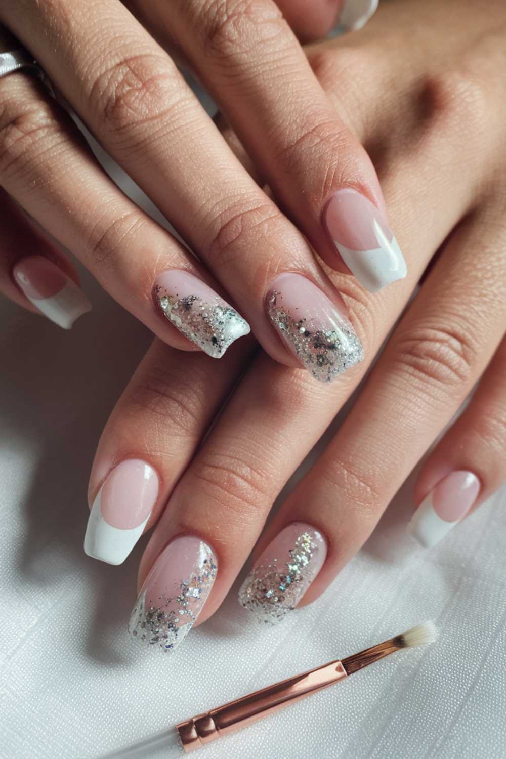 Close-up of hands with manicured nails. Two nails on each hand have silver glitter tips, while the others feature classic French tips. A small nail art brush lies nearby on a white surface.