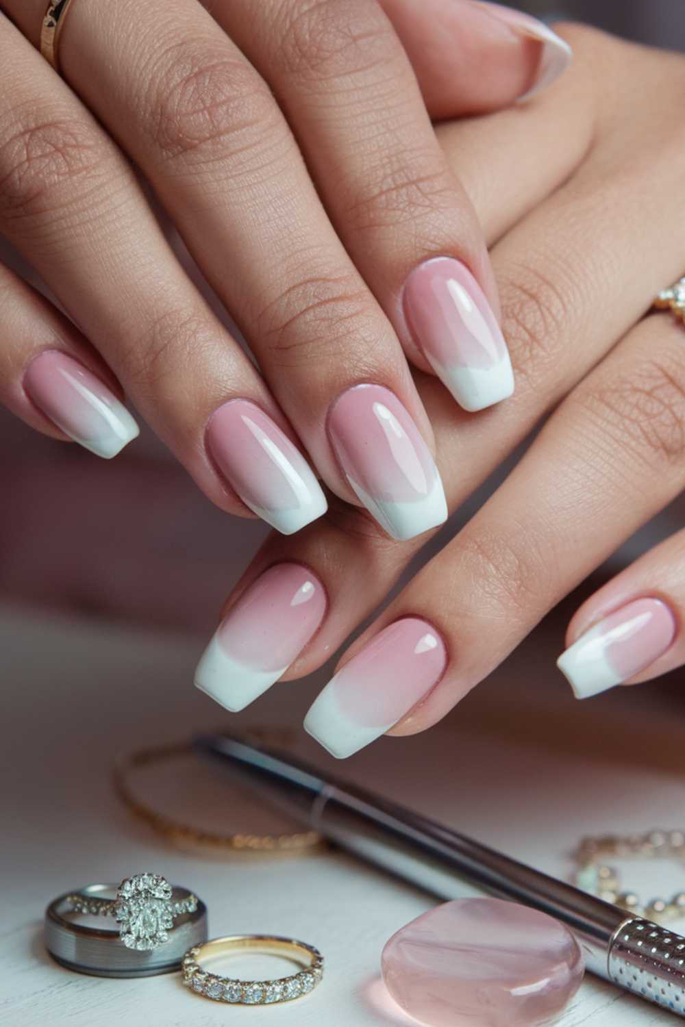 Close-up of hands with a French ombre manicure showcasing pink to white gradient nails. Jewelry, including rings and bracelets, is visible. A pen, a pink stone, and additional rings are placed nearby on a white surface.
