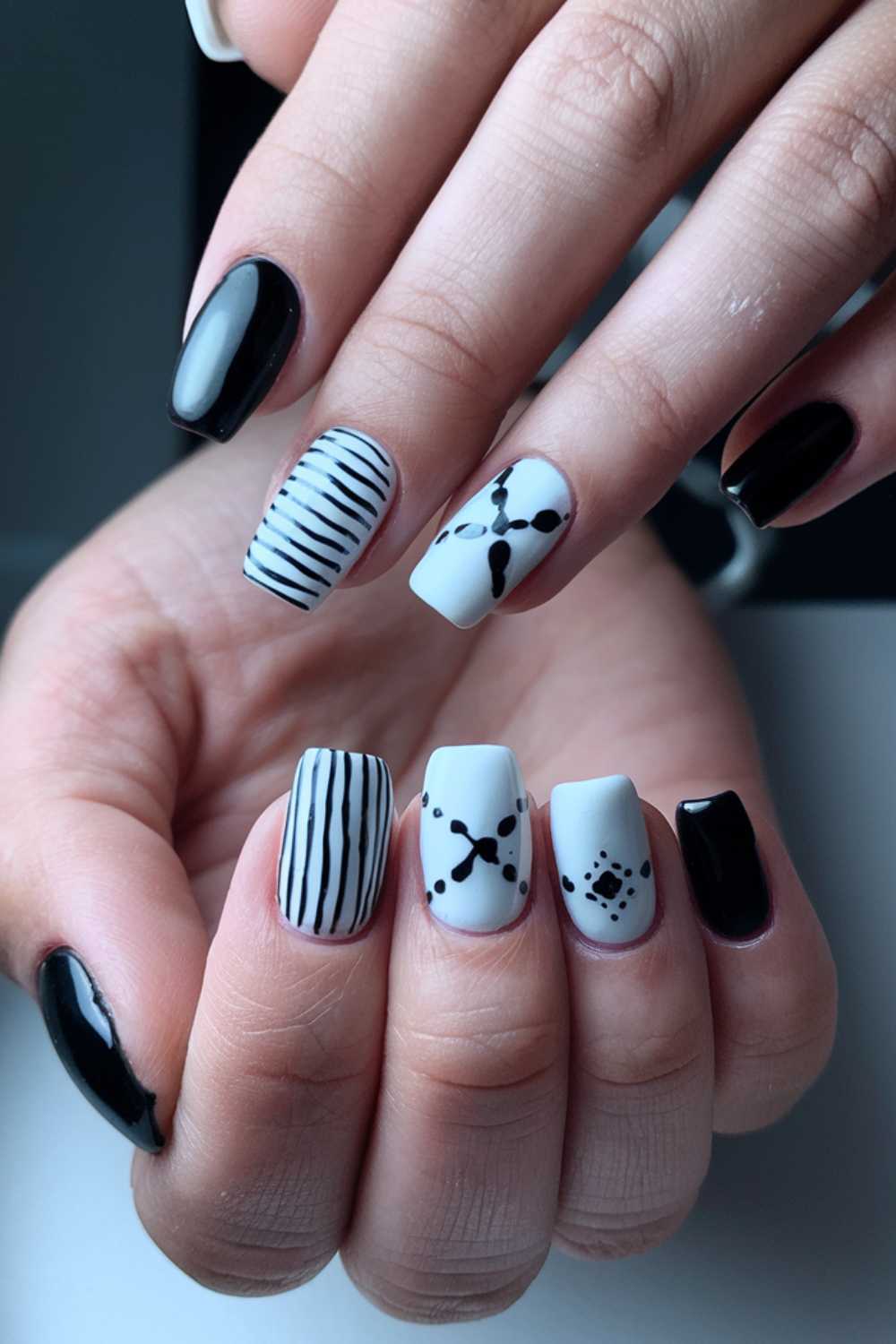 Close-up of a hand with manicured nails featuring various designs: black glossy nails, white nails with black stripes, and white nails with black abstract dotted patterns on a light blue background.