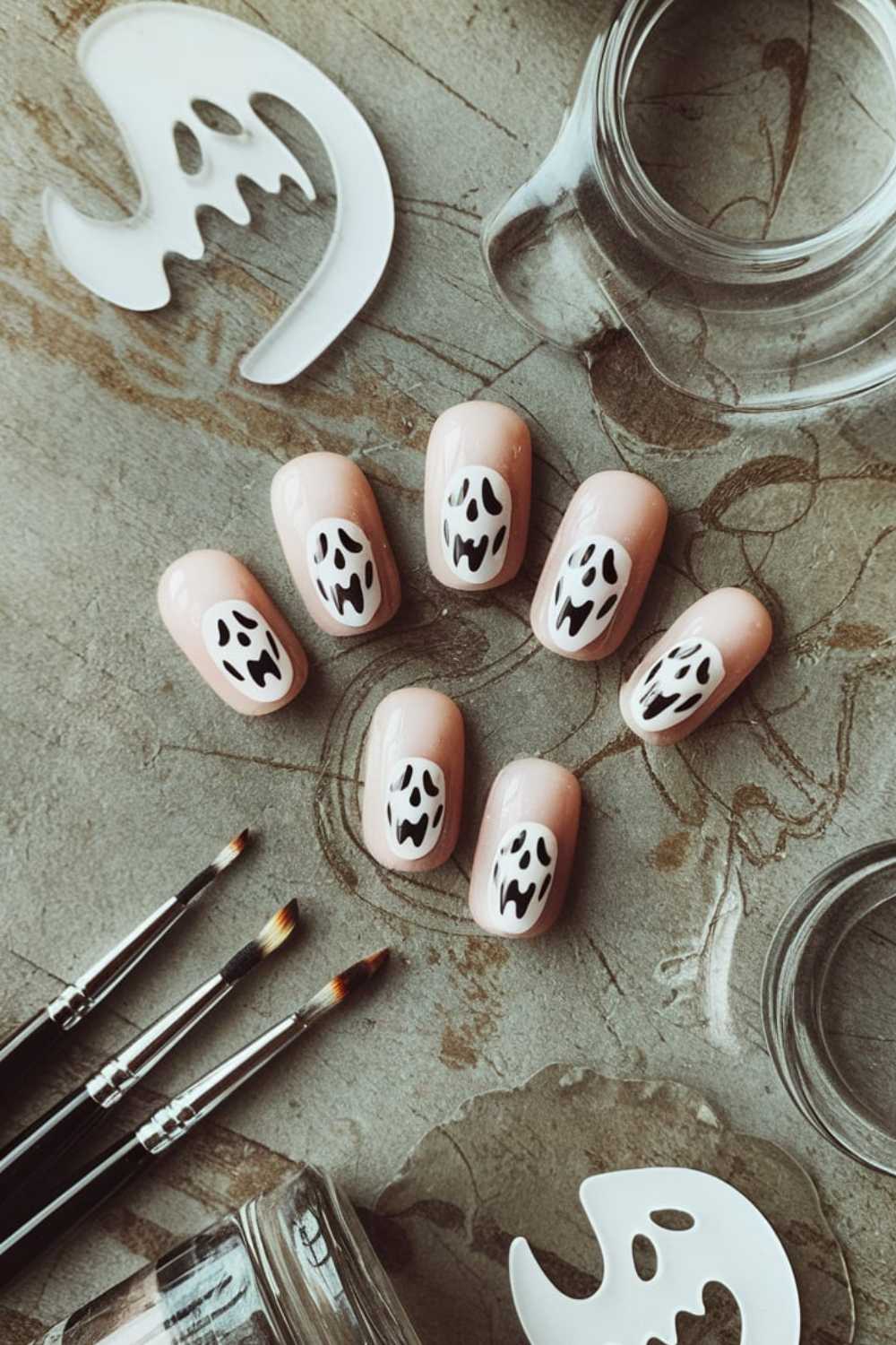 Nude-colored press-on nails with black and white ghost designs are arranged on a textured surface with transparent nail polish containers, brushes, and ghost-shaped decals nearby.