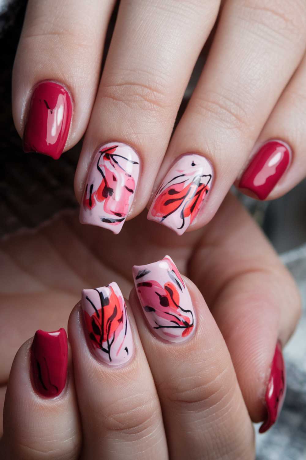 Close-up of hands showcasing cherry nail art designs. The nails feature a mix of solid red polish and pink polish with abstract red and black leaf-like patterns. The blurred background enhances the intricate details of the stunning nail art.
