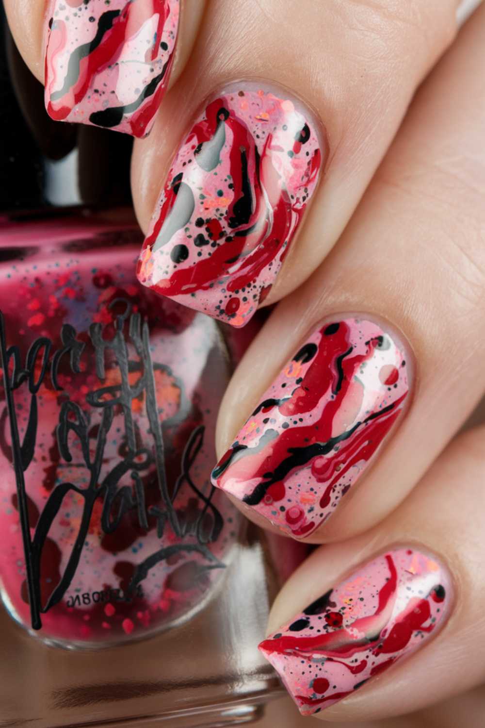 Close-up of a hand with vibrant, abstract cherry nail art designs. The nails boast red, pink, black, and white swirls with speckled patterns resembling splattered paint. A matching nail polish bottle is showcased in the background.