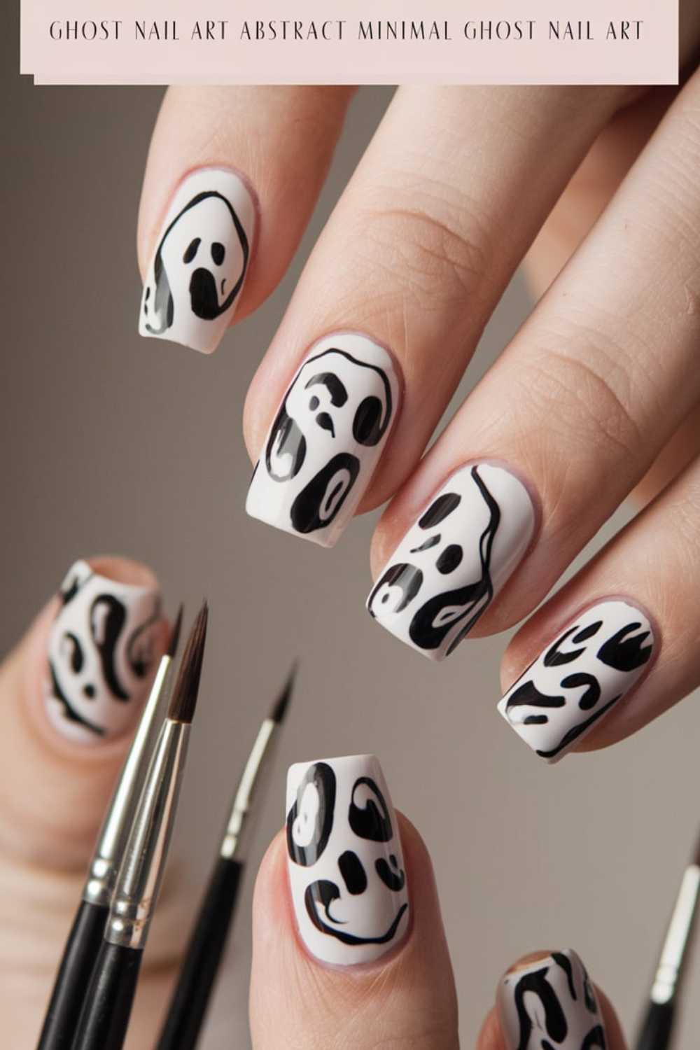 Close-up of a hand with nails painted in black and white ghost-themed designs. The background is neutral, and there are paintbrushes visible beside the hand. The text at the top reads, Ghost Nail Art Abstract Minimal Ghost Nail Art.