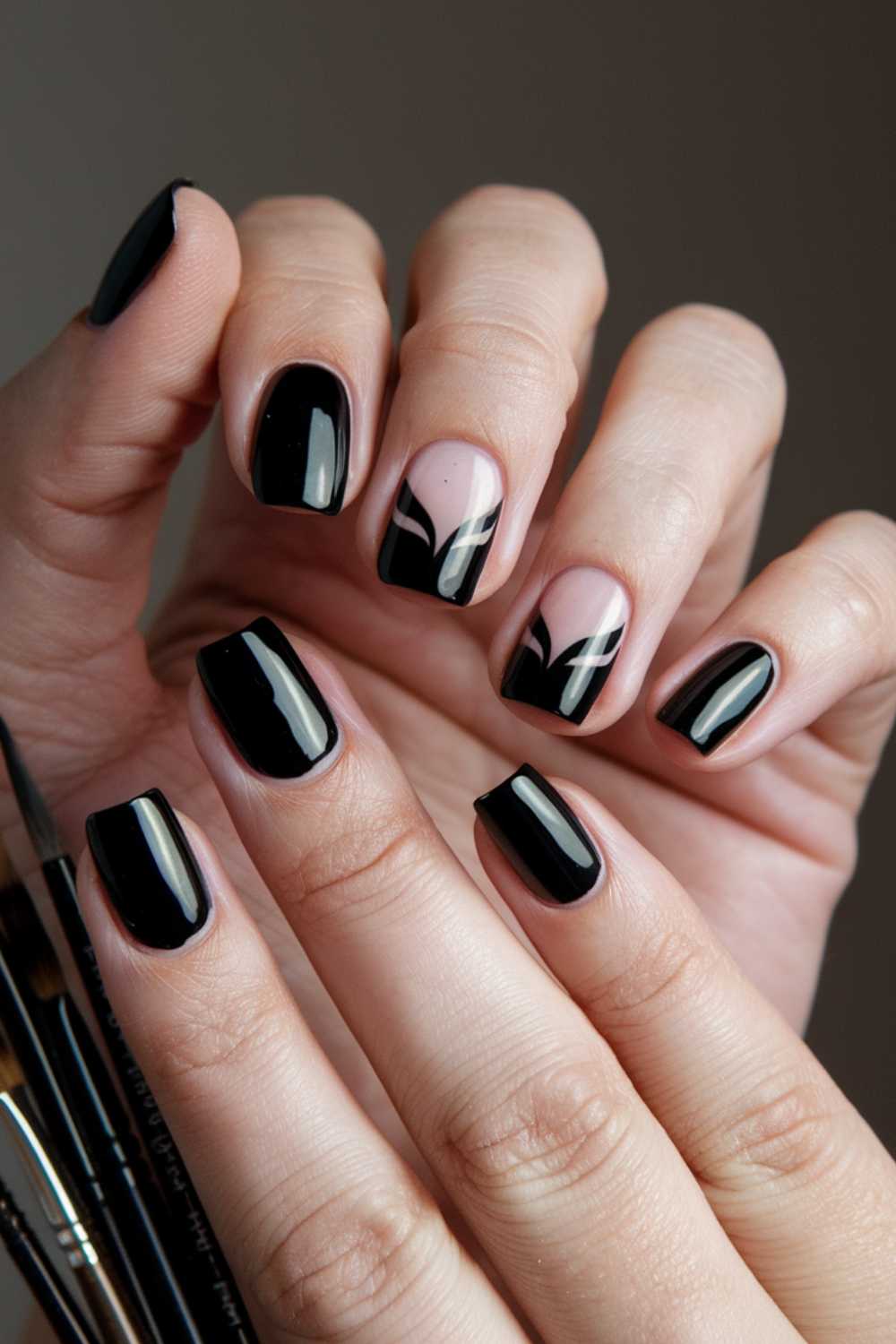 Close-up of a hand with manicured nails. Four nails are painted glossy black, while one has a black and white geometric pattern on a nude base. Brushes are visible in the background.