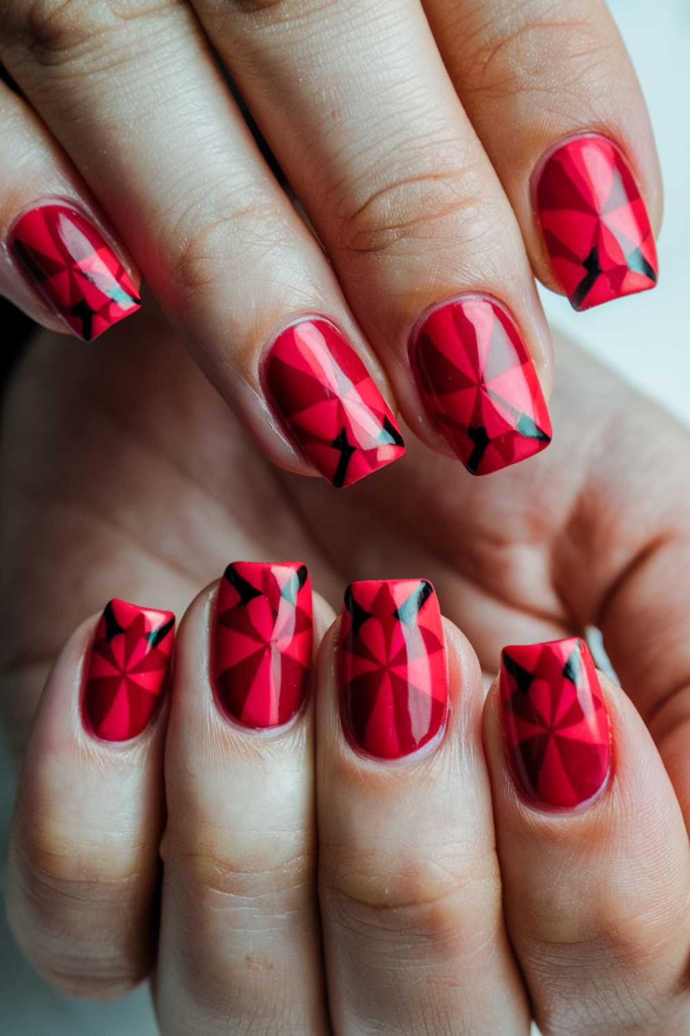 Close-up of hands with manicured nails featuring a geometric design. The nails are painted red with black angular patterns, creating a striking contrast. The nails are neatly trimmed and glossy, showcasing a modern and artistic look.