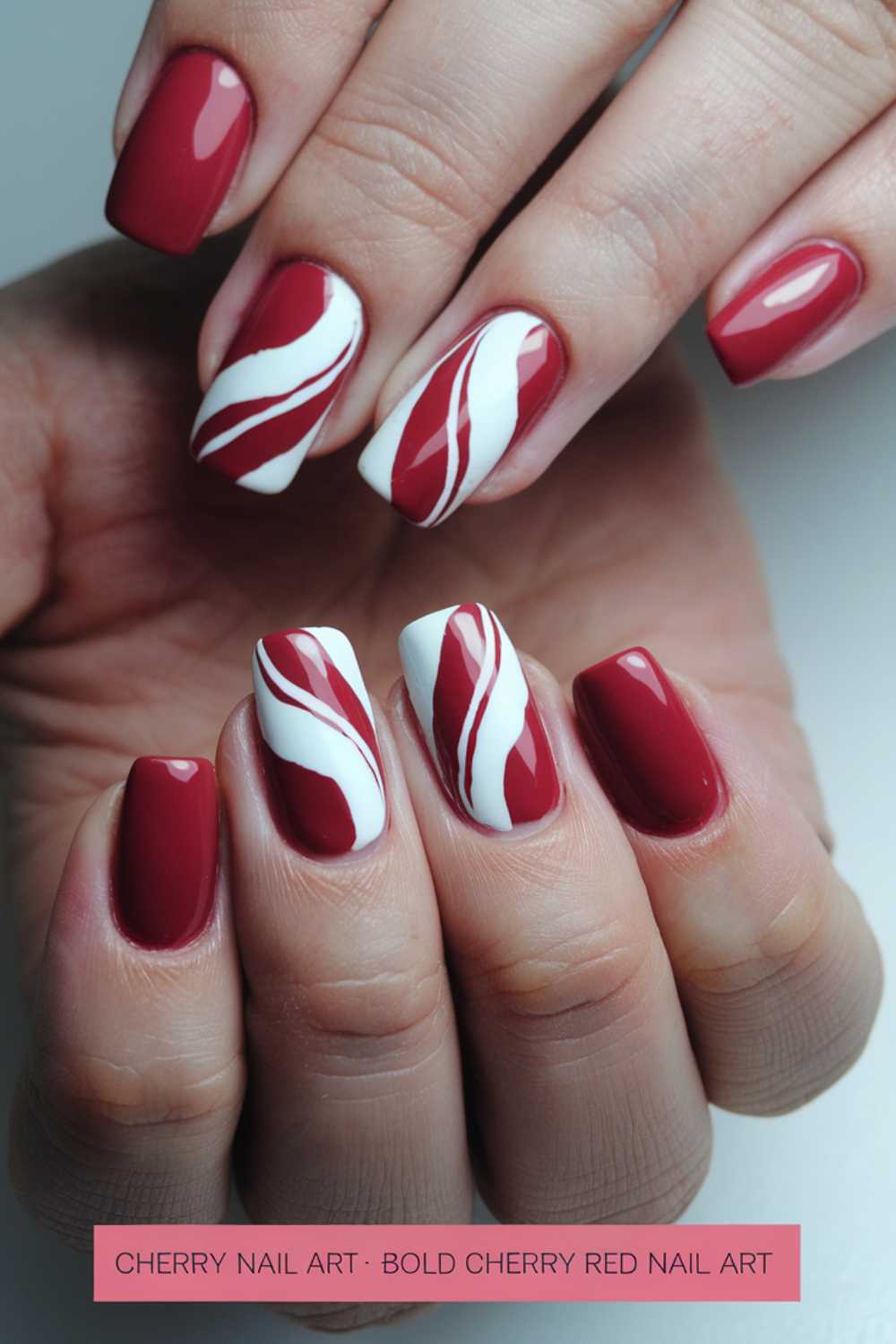 Close-up of hands with manicured nails showcasing cherry nail art designs. Nails are painted in cherry red and white, featuring bold, flowing wave patterns. The design alternates between solid red and red with white swirl accents for a dynamic look.