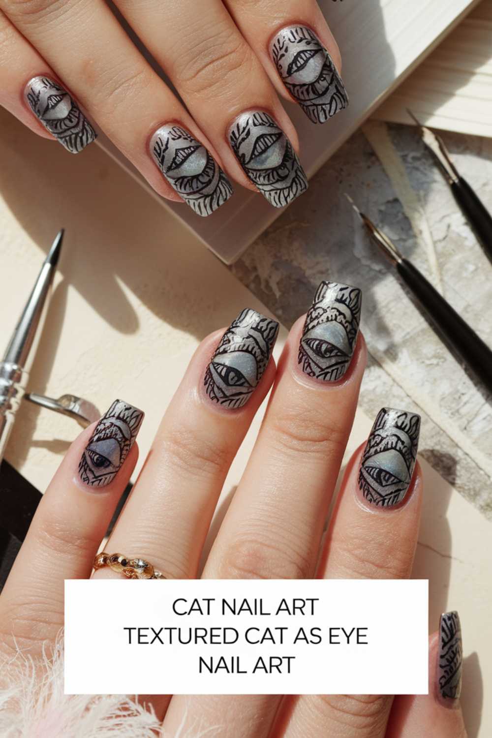 Close-up of hands showing nails with intricate cat eye designs. Each nail features a stylized eye with fur-like textures surrounding it. Art tools are visible in the background, suggesting a nail art setup.