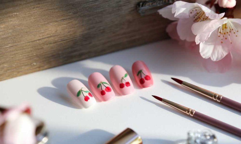 A set of artificial nails featuring cherry nail art designs in pink and red is elegantly displayed on a white surface. Nearby, two nail art brushes accompany the scene, enhanced by pink flowers and makeup items that create a charming backdrop.
