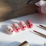 A set of artificial nails featuring cherry nail art designs in pink and red is elegantly displayed on a white surface. Nearby, two nail art brushes accompany the scene, enhanced by pink flowers and makeup items that create a charming backdrop.