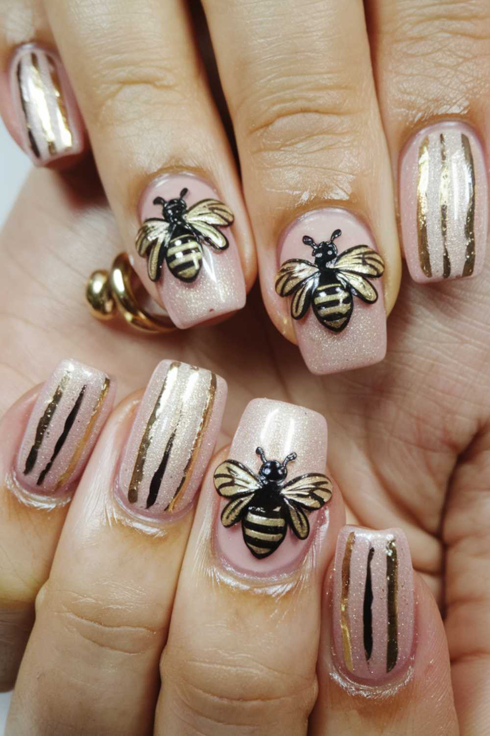 Close-up of a hand with manicured nails in shimmering light pink. Each nail showcases vertical gold stripes, while some boast a dazzling 3D bee nail art decoration, intricately detailed in black and gold.