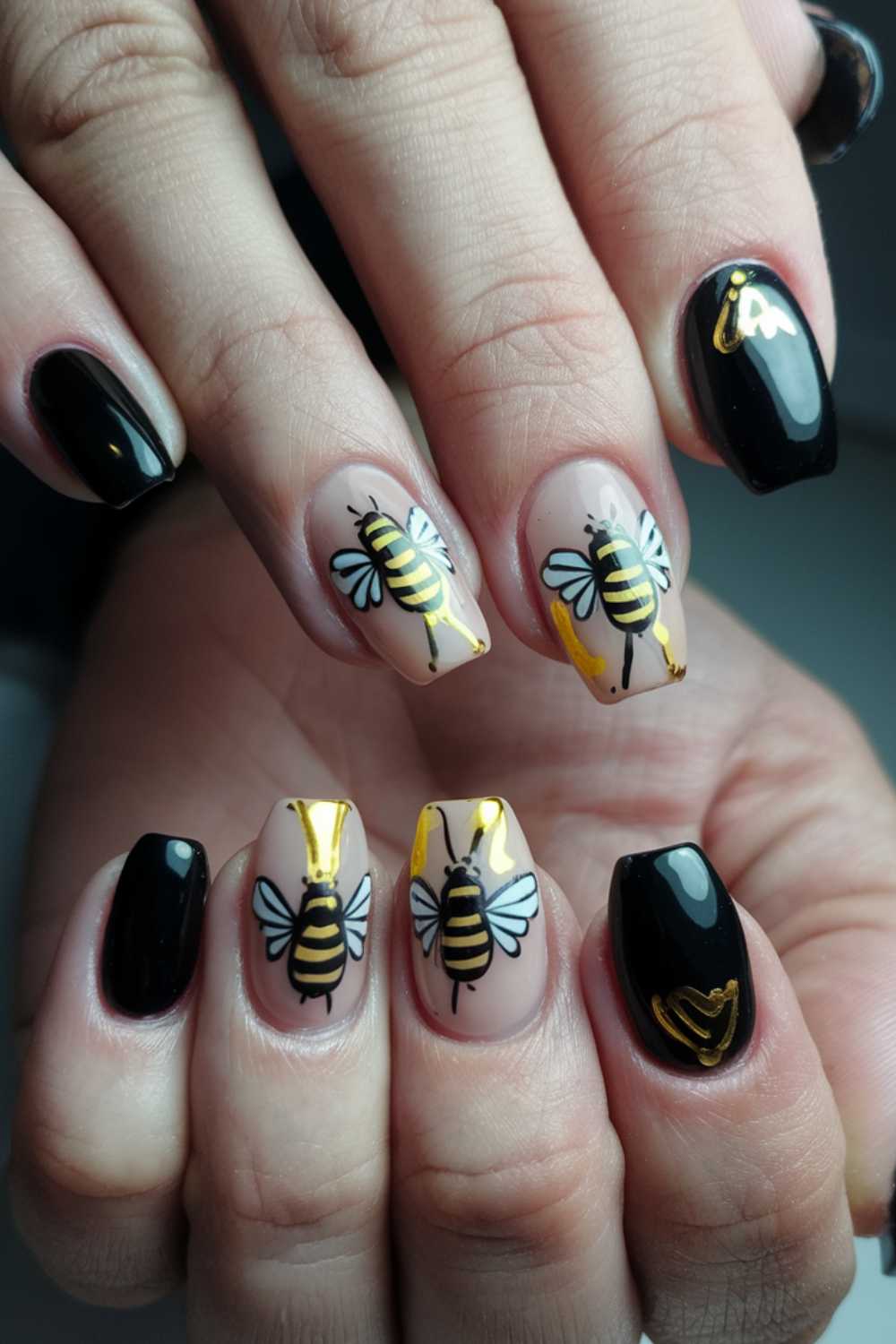 A close-up of hands showcases stunning bee-themed nail art. This bee nail art features alternating glossy black and clear nails adorned with intricate bee designs, complete with yellow and black stripes, delicate wings, and shimmering gold accents.