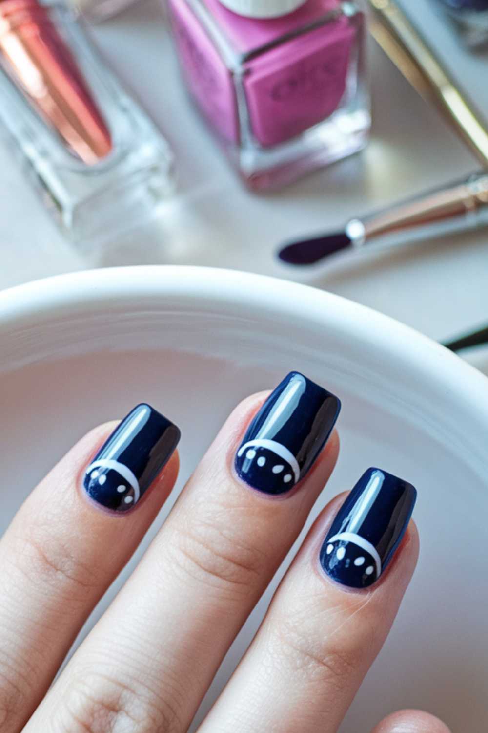 Close-up of a hand with dark blue nail polish and white crescent moon details on the nails. In the background, there are bottles of nail polish in various colors and a small paintbrush.