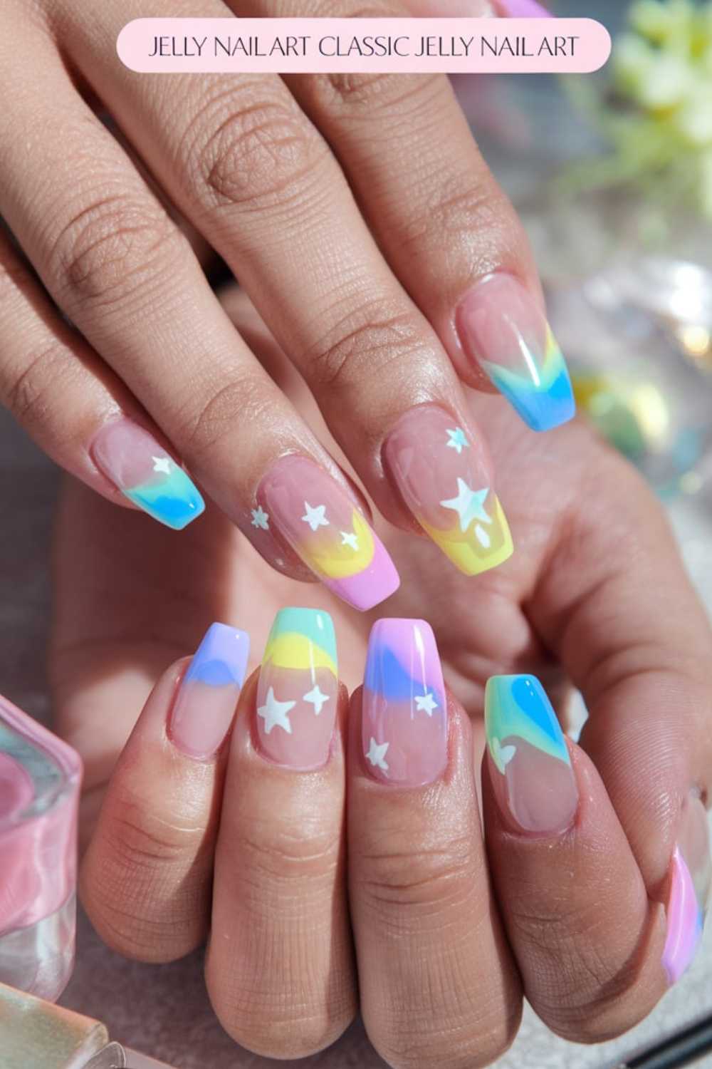 Close-up of hands showcasing vibrant jelly nail art ideas with a gradient of pink, yellow, blue, and green tips. White star stickers adorn the nails against a softly blurred background, hinting at flowers and a product bottle.