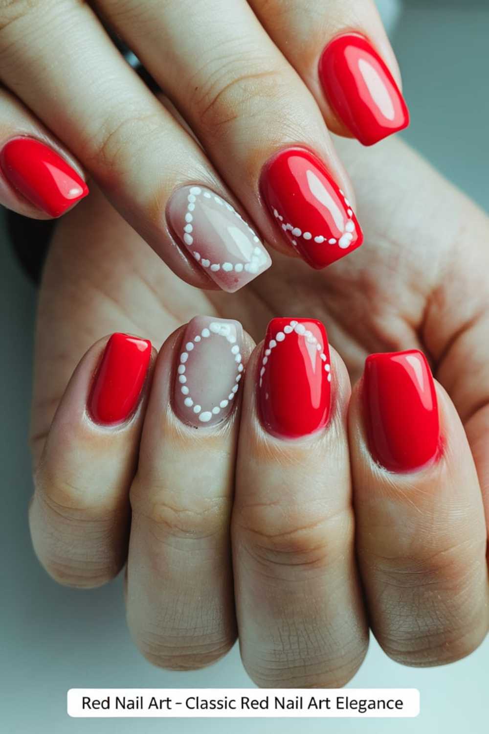 Close-up of hands with a manicure. Fingers show a mix of glossy red nails and one nail on each hand painted nude with delicate white dot patterns.