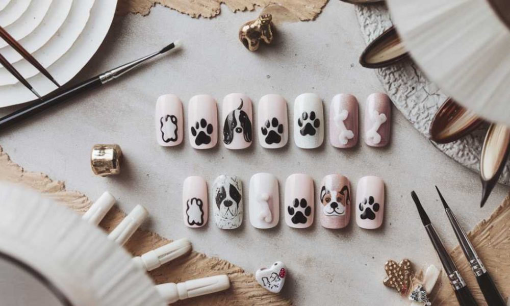 A set of false nails adorned with dog nail art, featuring cute paw prints and dog faces, arranged on a textured surface. Surrounding the nails are various nail art tools and decorative items.