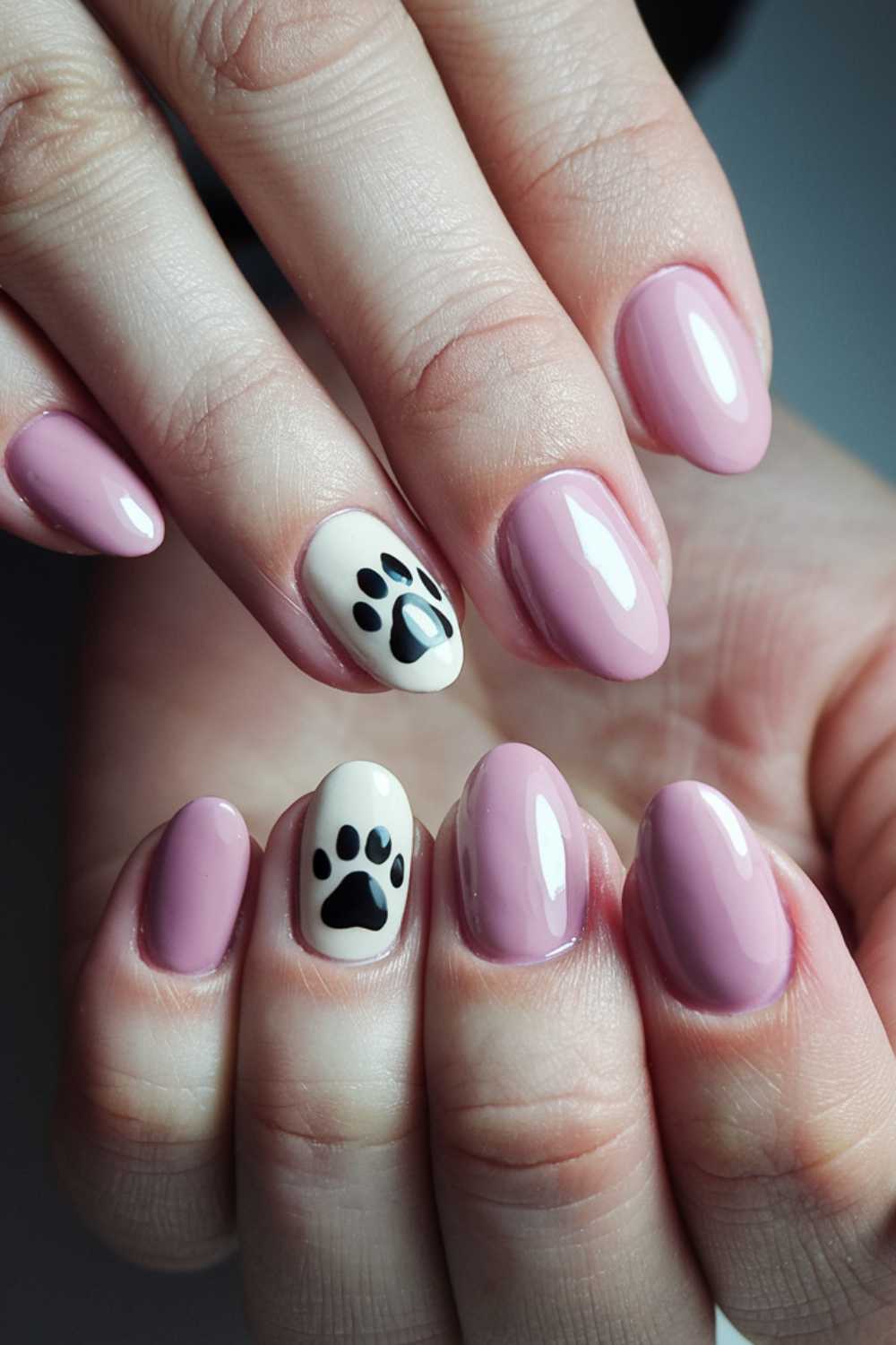 Close-up of hands displaying a manicure with glossy light pink nails. Embracing the dog nail art trend, one nail on each hand is painted white and features a black paw print design. The nails are shaped in a natural oval contour.