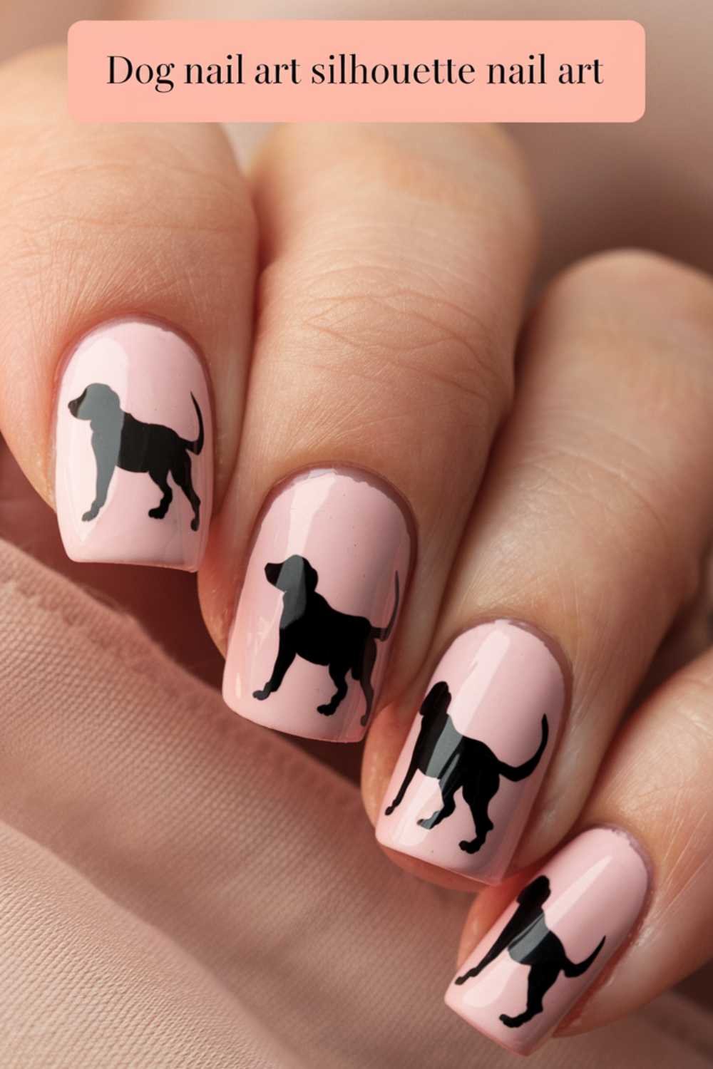 Close-up of nails with light pink polish adorned with black dog silhouettes. Dog Nail Art elegantly graces the top, as the nails rest gently on soft pink fabric.