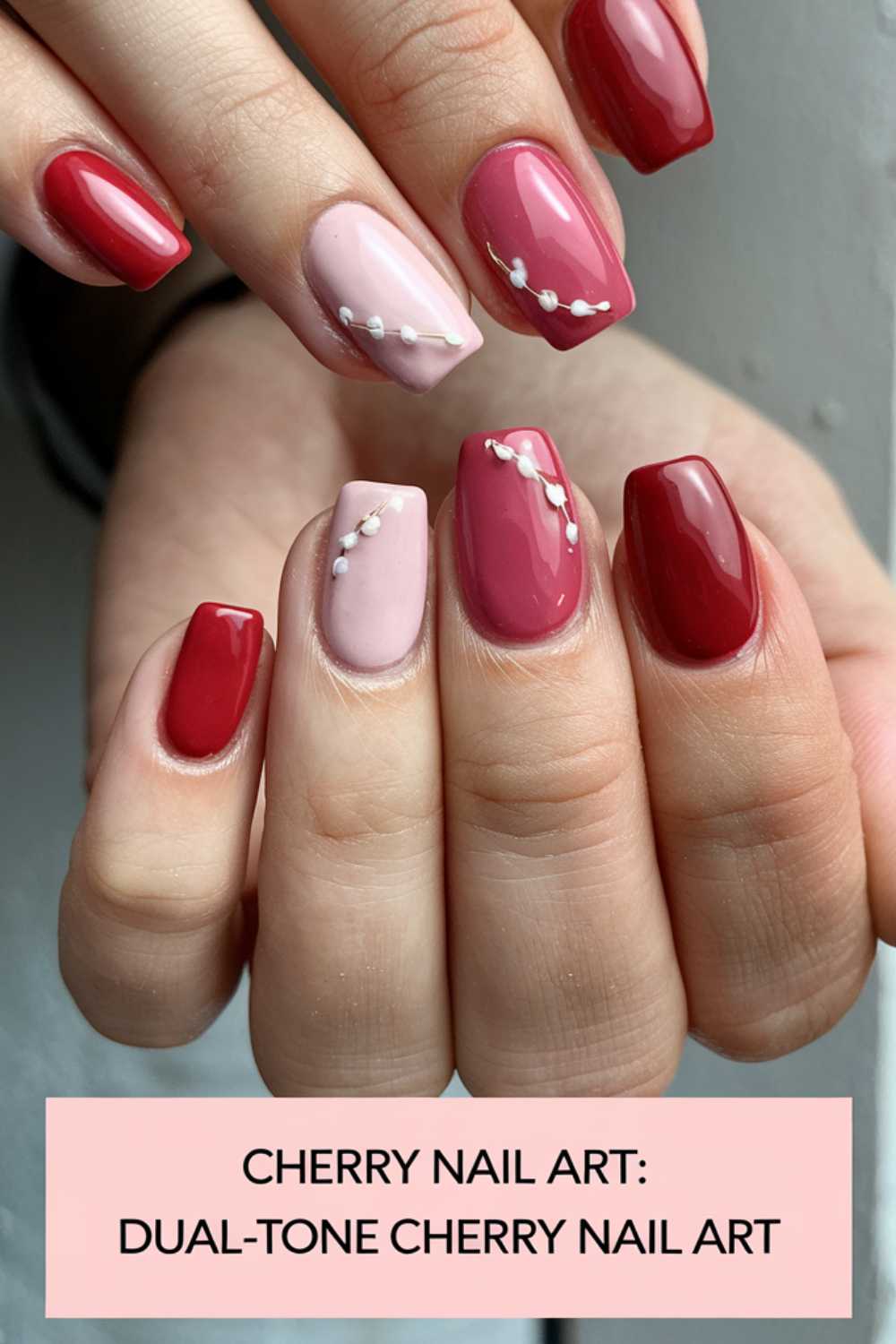 Close-up of hands with cherry-themed nail art. Nails feature dual-tone red and pink shades with small white bead accents. Text at the bottom reads: Cherry Nail Art Designs: Dual-Tone Cherry Nail Art.