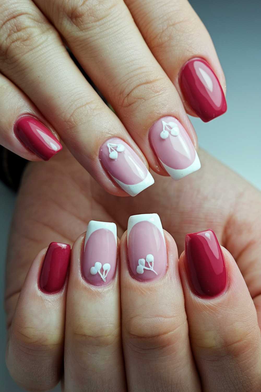 Close-up of hands showcasing manicured fingernails. The nails boast glossy red polish and a chic pink French manicure with white tips. Some pink nails are beautifully adorned with cherry nail art designs, giving the look an elegant and stylish flair.