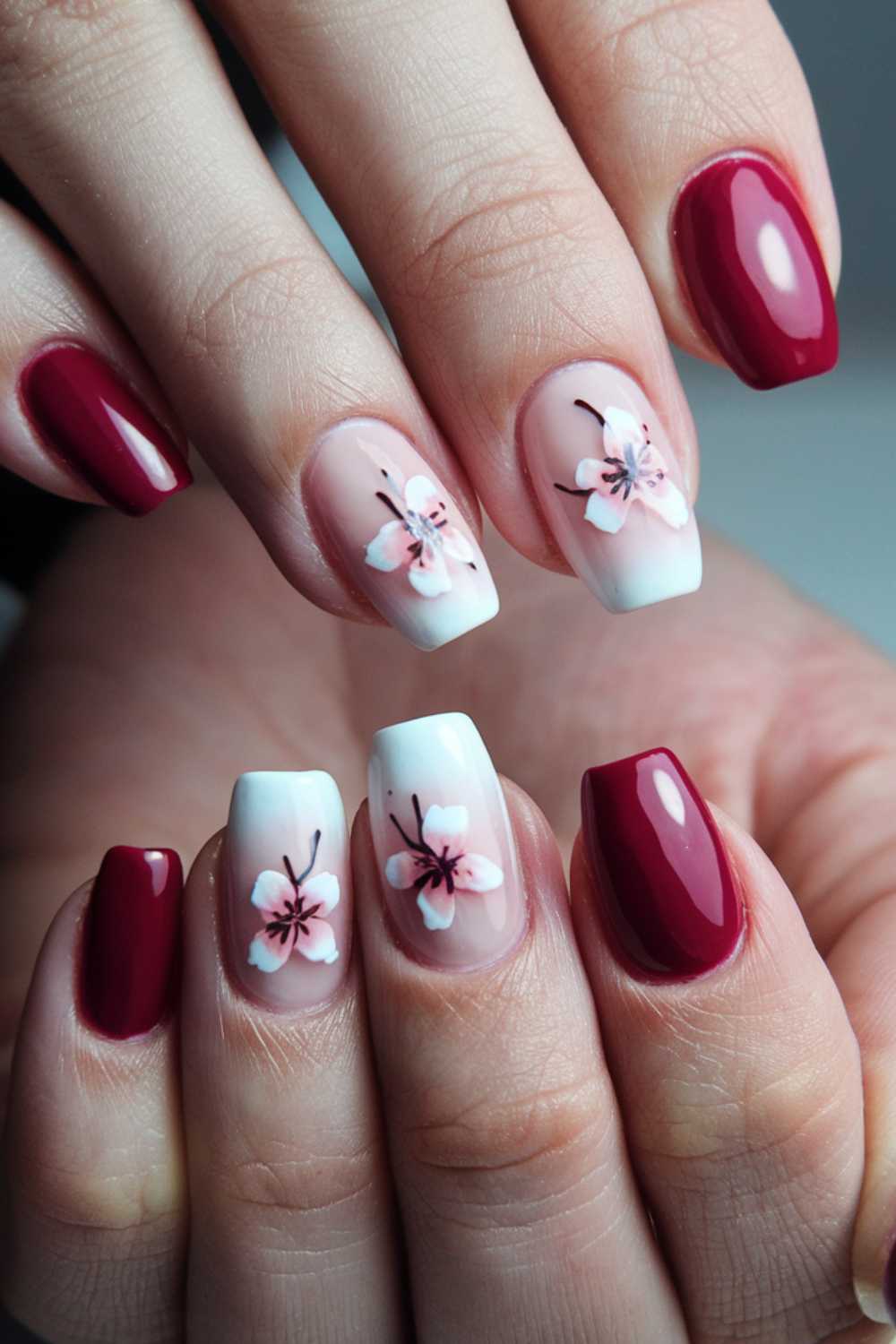 A close-up of hands with manicured nails showcases a stunning display of cherry nail art designs. Four nails boast a glossy red polish, while four feature pink flowers with black centers on a white background. The middle nails seamlessly gradient from white to natural, enhancing the floral theme.