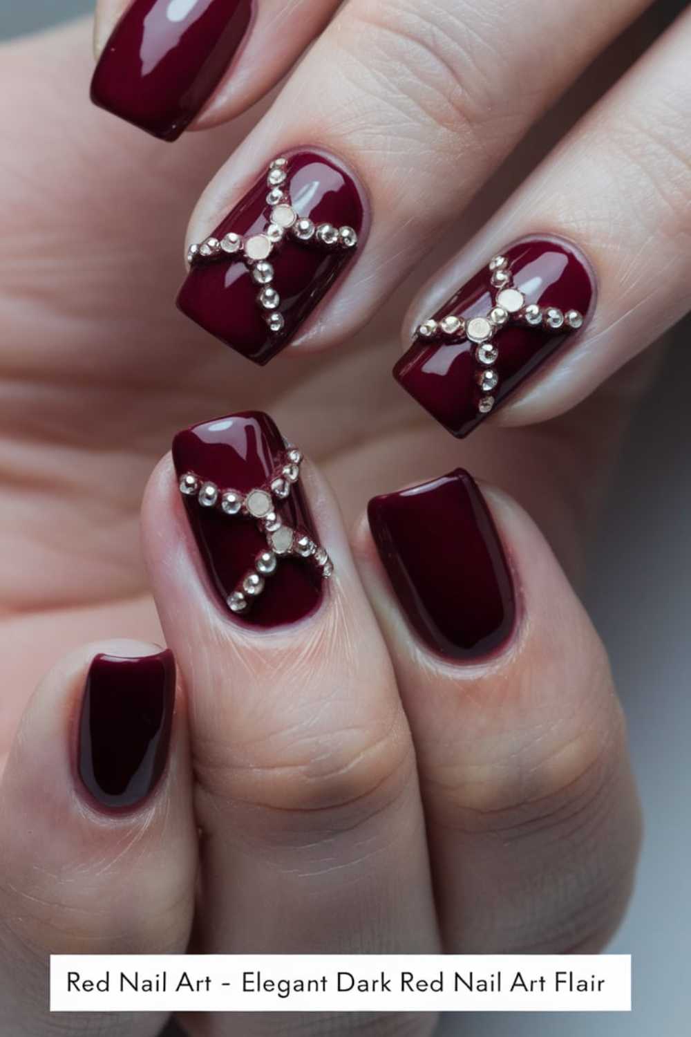 Close-up of hands with dark red manicured nails featuring metallic bead accents in a crisscross pattern on some nails. The nails are glossy and elegantly styled.