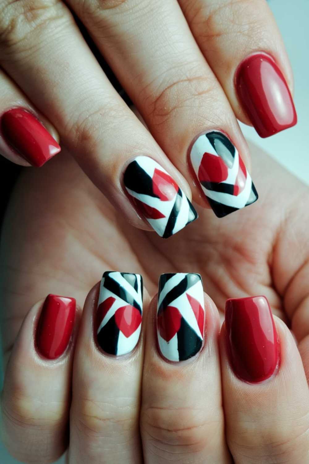 Close-up of hands with a manicure showcasing cherry nail art designs. Nails boast a glossy finish, blending red, black, and white hues. Some nails feature geometric patterns in red and black on a pristine white backdrop.