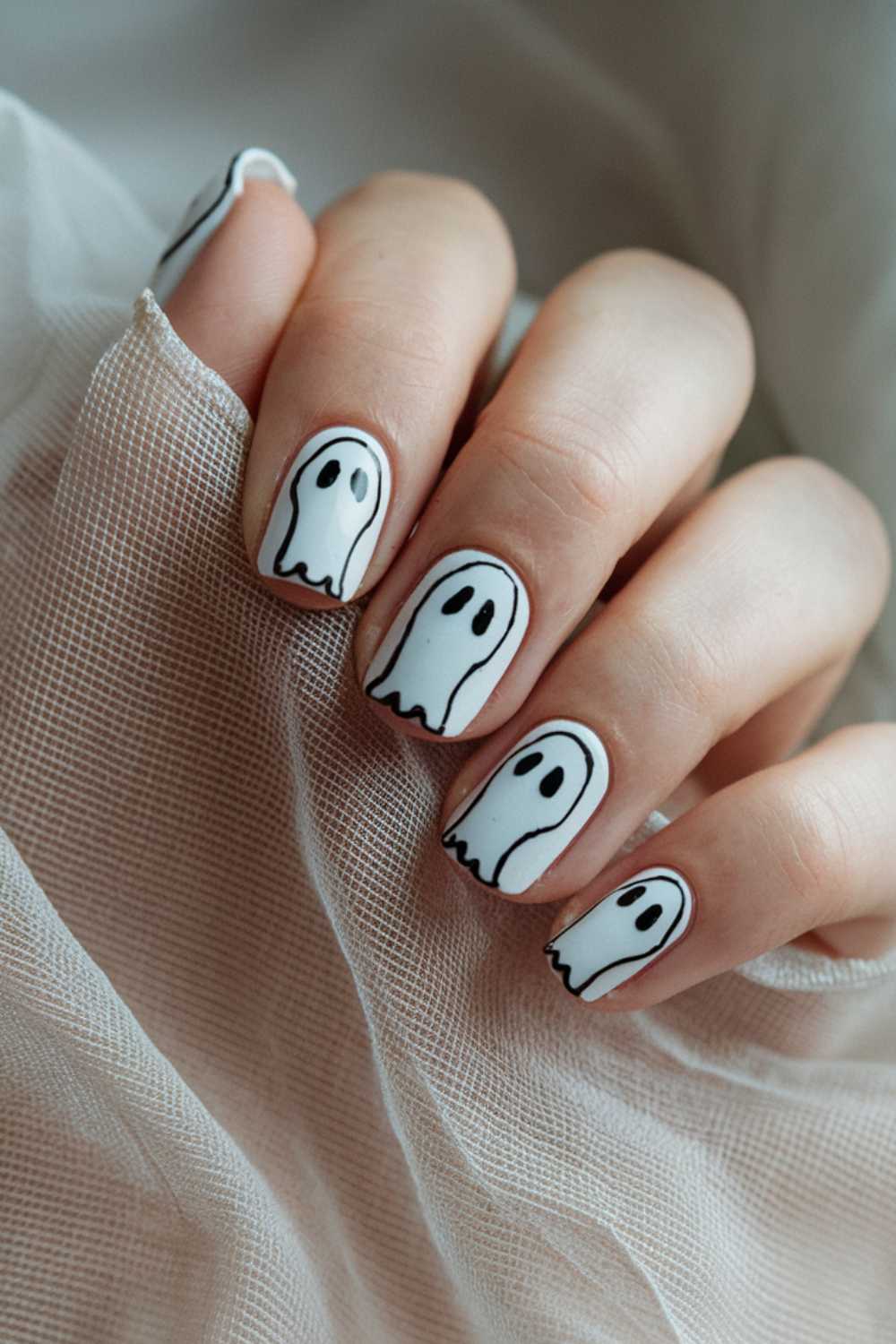 A hand with nails painted white features cute ghost designs, each nail decorated with a simple black-outlined ghost. The hand rests gently on a sheer white fabric, creating a soft and spooky vibe.