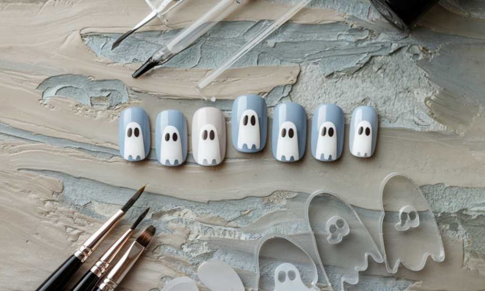 Artificial nails painted with ghost faces in shades of blue and white are arranged on a textured surface. Painting tools, including brushes and clear ghost-shaped nail templates, surround the nails.