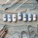 Artificial nails painted with ghost faces in shades of blue and white are arranged on a textured surface. Painting tools, including brushes and clear ghost-shaped nail templates, surround the nails.