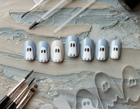 Artificial nails painted with ghost faces in shades of blue and white are arranged on a textured surface. Painting tools, including brushes and clear ghost-shaped nail templates, surround the nails.