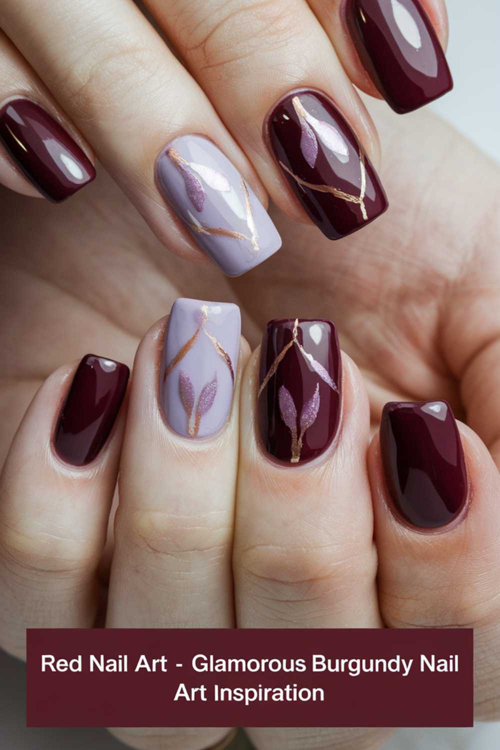 Close-up of a hand with glossy burgundy and gray nail art. The design features metallic accents and geometric patterns on the middle and ring fingernails. The colors and shine create a sophisticated, elegant look.