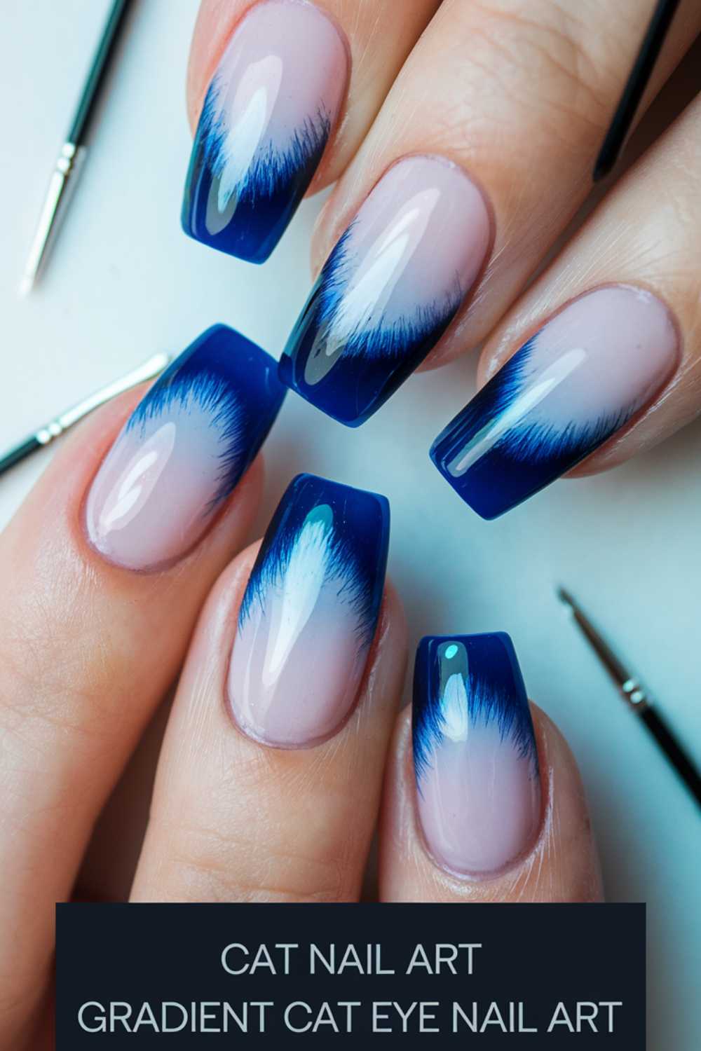 Close-up of hands showcasing nail art. The nails have a gradient cat eye design with a blend of light pink at the base transitioning to deep blue tips. Thin brushes are also visible next to the hands.