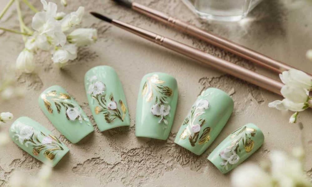A set of mint green artificial nails featuring white flowers and gold leaf designs, arranged beside two nail art brushes and surrounded by delicate white flowers, perfect for showcasing elegant green nail art.